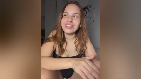 Video of a young, smiling woman with light skin and long, wavy brown hair, wearing a black bra, sitting indoors.