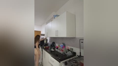 Video of a slender, nude woman with long brown hair, standing in a small, cluttered kitchen with white cabinets and a black countertop, holding a red object.