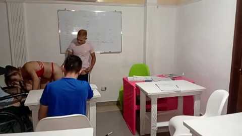 Media: Video of a small, white-walled classroom with two men and a woman in red lingerie on the floor; one man wearing a blue shirt and another in a pink shirt, white tables, green chairs, and a whiteboard in the background.