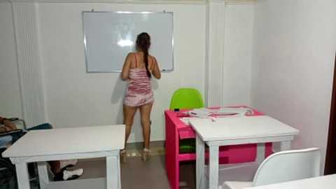 Video of a young woman with long brown hair, wearing a pink camisole and shorts, standing in a classroom with white walls, a whiteboard, and pink and green tables.