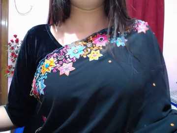 Video of a woman with long black hair, wearing a black velvet shirt with colorful embroidered stars and flowers, indoors with red curtains and floral arrangement in the background.