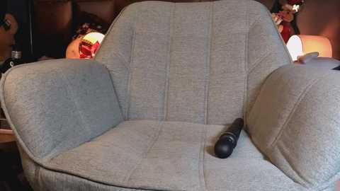 Video of a cozy, mid-century modern armchair with a gray fabric seat and backrest, featuring a black sex toy on the armrest. The background shows a dimly lit room with warm lighting and decorative elements, including a red candle and a white lampshade.