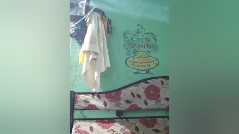 Video of a green wall with a cartoonish alien character, a white towel hanging, and a floral-patterned bedspread with red and pink flowers.