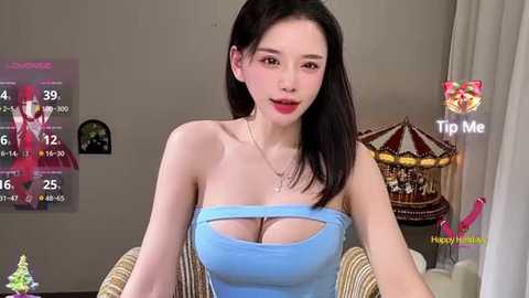 A video of an East Asian woman with long black hair and light skin, wearing a strapless blue top that reveals her cleavage. She sits in a wicker chair in a cozy room with a festive atmosphere.