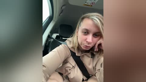 Media: Video of a blonde woman with shoulder-length hair, wearing a beige uniform with a black shoulder strap, sitting in a car, resting her chin on her hand, with a blurred background.
