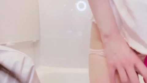 A close-up video of a fair-skinned hand in a white, possibly sheer, garment, partially submerged in a bathtub, with a blurred, white-tiled bathroom background.