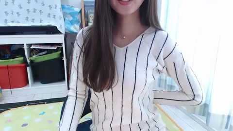 Video of a smiling young woman with long brown hair, wearing a white and black striped sweater, sitting on a polka-dotted mat in a brightly lit room with shelves, baskets, and toys.