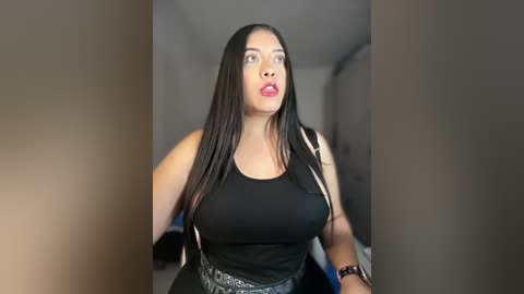 Video of a plus-size woman with long, straight black hair, wearing a black tank top and high-waisted jeans. She has a medium complexion and wears bright red lipstick. The background is a blurred indoor space with neutral tones.
