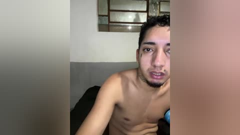 Video of a young, shirtless Asian man with short dark hair and a trimmed beard, partially hidden by a large, blurred object. Background includes a white wall with a rectangular mirror.