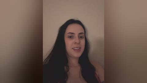 Video of a young woman with long black hair and fair skin, standing in a simple, beige-walled room. She has a neutral expression and is wearing a dark top.