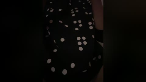 A close-up video of a dark fabric with white polka dots, possibly a dress or skirt, dimly lit, with shadows suggesting a person wearing it.