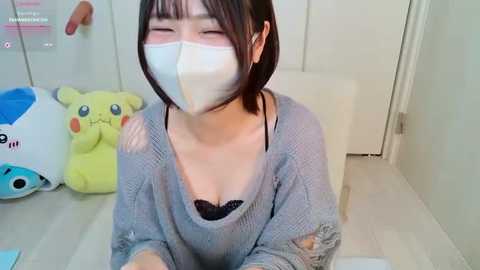 Video of an Asian woman with short dark hair, wearing a white face mask and a light gray sweater over a black lace bra, sitting on a white chair in a white room with a Pikachu plush toy in the background.