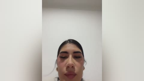 A video of a young woman with dark hair and light skin, wearing makeup, standing in a minimalist, white-walled room.