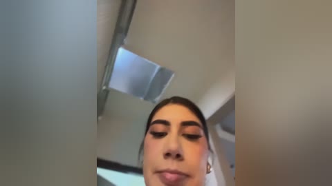 A close-up video of a woman with light skin, brown hair, and subtle makeup, including mascara and blush, gazing upward. Background shows a modern, minimalist ceiling with a recessed light fixture.