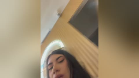 A blurred video of a young woman with long dark hair, wearing makeup and a black top, leaning against a beige wall with a small mirror reflection.