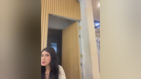 Video of a young woman with long black hair, light skin, and a slender build, standing in a modern hallway with beige walls and a textured yellow door. She has a neutral expression.