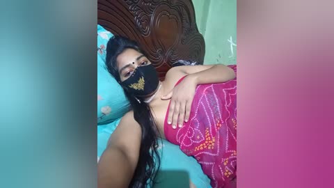 Video of a woman with long black hair, wearing a black face mask and a red sari with floral patterns, lying on a teal bedspread, with a wooden headboard in the background.