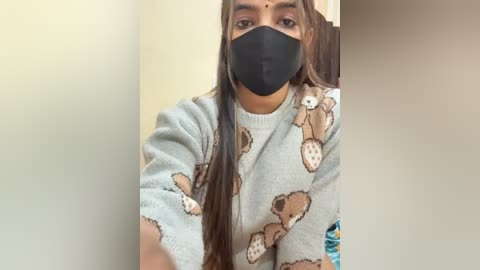 Video of a young woman with long, straight brown hair, wearing a gray sweatshirt with teddy bear prints, a black face mask, and sitting indoors with a beige wall background.