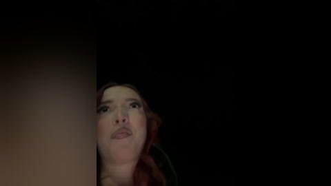 Video of a young woman with fair skin, long brown hair, and full lips, gazing upward with a contemplative expression against a dark, gradient background.