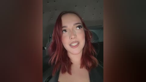Video of a young woman with shoulder-length, wavy, reddish-brown hair, fair skin, and full lips, smiling. She is wearing a black top. The background features a dimly lit, plush interior with small, circular lights.