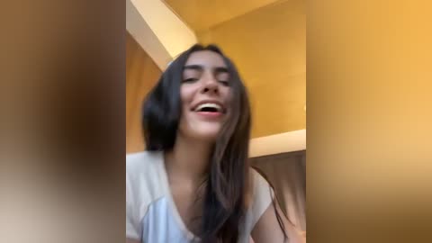 Media: A video of a young woman with long dark hair, smiling widely, wearing a light-colored shirt, standing in a modern, yellow-tiled room with a white counter.