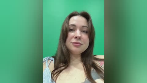 Video of a young Caucasian woman with long brown hair, fair skin, and a slight smile, wearing a light blue, patterned top. Green screen background.
