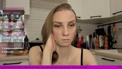 Video of a blonde woman with a serious expression in a modern kitchen, wearing a black top, with a digital overlay showing her weight and fitness stats.