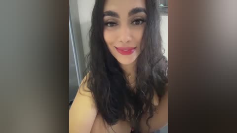 Video of a woman with long, wavy black hair, wearing a yellow top, standing indoors with blurred background, smiling with red lipstick.