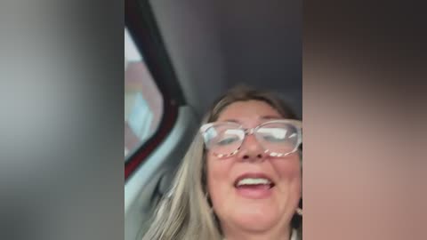 Media: A video of a smiling woman with long blonde hair and glasses, partially visible, taken from inside a car with a blurred window and car interior visible in the background.