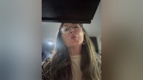 Video of a woman with glasses and long brown hair, wearing a beige top, peeking from under a dark wooden table, slightly blurred background.