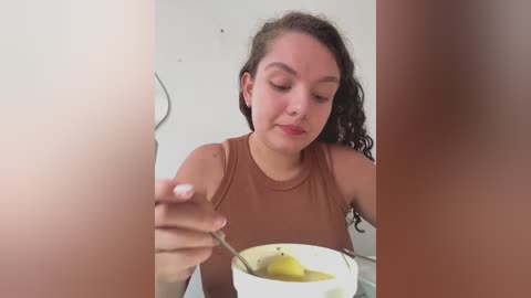 Video of a curly-haired woman with light skin in a brown tank top, holding a white bowl with a yellow substance, partially obscured by a blurry vertical object.