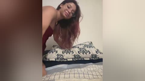 Video of a smiling woman with long, wavy, dark red hair, wearing a red top, bending over a bed with black and white patterned sheets.