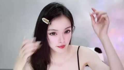 Video of an East Asian woman with long, straight black hair styled with a gold hair clip, wearing a black spaghetti strap top, smiling. Soft-focus background with white and pink hues.