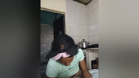 Media: Video of a woman in a mint-green shirt, dark hair, and pink hat, bending over in a small, tiled kitchen with white walls and a silver pot on the stove.
