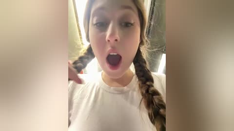 Media: Video of a young woman with light skin, brown hair in braids, wearing a white t-shirt, open-mouthed, in a dimly lit room with blurred background.