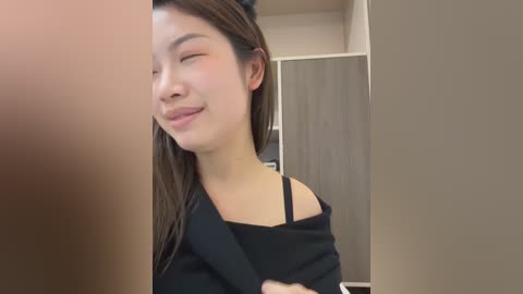 Video of a young East Asian woman with long brown hair, wearing a black off-shoulder top, smiling and winking in a modern, beige-walled room with a wooden door in the background.
