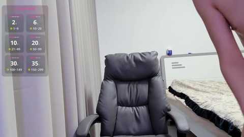 A video captures a nude woman with fair skin, standing beside a gray leather office chair in a minimalist bedroom with white walls, beige curtains, and a neatly made bed.