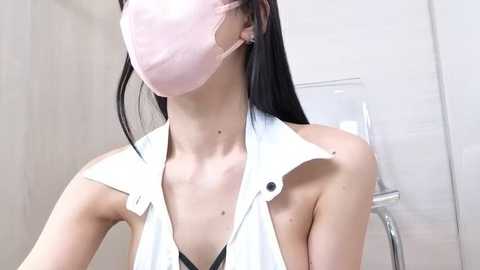Video of a slender, light-skinned woman with long black hair, wearing a pink face mask and a white, open-fronted, sleeveless shirt, standing in a modern, white-tiled bathroom.