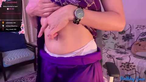 Video of a woman with light skin, wearing a purple top, white panties, and a watch, lifting her shirt to show her midriff in a bedroom with floral decor and a chair.