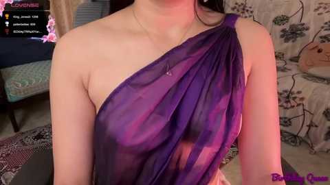 A video of a woman with light skin wearing a sheer, purple one-shoulder dress, indoors with floral-patterned wallpaper and a TV in the background.