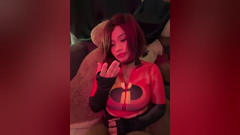 Video of a young woman in a red, tight Incredibles costume, sitting on a plush teddy bear, indoors with a red filter, dim lighting.