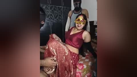 A video captures a woman in traditional Indian attire, adorned with gold and red makeup, seated on a bed. She wears a red blouse and long skirt, with a man standing behind her. The background features dark, patterned wallpaper.