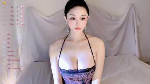 A video of an Asian woman with fair skin, dark hair tied back, wearing a revealing leopard print top that emphasizes her ample cleavage. She sits on a bed with white sheets, draped in sheer white fabric, in a dimly lit room.