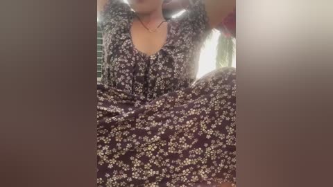 A video of a woman with medium skin tone, wearing a purple floral dress, sitting with her legs spread. She has a slender build and is indoors, with soft, diffused lighting.