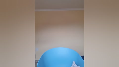 Video of a minimalist, beige-walled room with a large, round, bright blue exercise ball in the center. The ball casts a shadow on the floor, suggesting movement. The scene is simple and uncluttered.