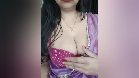 A close-up video of a South Asian woman with fair skin, long black hair, and full lips, wearing a purple satin blouse with a pink polka-dotted bra, adjusting her bra strap.