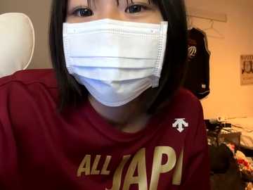 Video of a woman with short black hair, wearing a white surgical mask, maroon All-American University jersey, indoors with cluttered background.