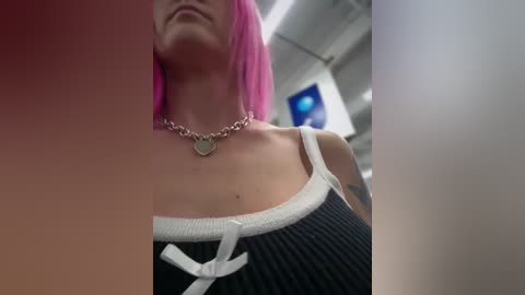 Video of a woman with bright pink hair, wearing a black ribbed tank top with white trim and a small bow, silver necklace, and visible collarbone. Background shows an airplane interior with blue lighting and overhead compartments.
