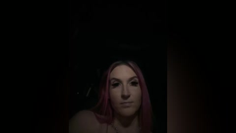 Video of a young woman with long, straight pink hair, fair skin, and a slight smile, set against a dark, shadowy background.