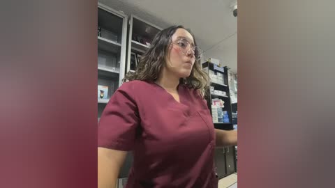 Media: Video of a middle-aged woman with wavy, shoulder-length hair, wearing glasses, and a maroon scrub top, standing in a pharmacy aisle with shelves of medicine and products.